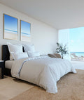 Ein modern hergerichtetes Bett mit einem glatten, weißen Excellence Bettdeckenbezug in einem luxuriösen, hellen Schlafzimmer mit großer Fensterfront und Blick auf den sonnigen Sandstrand.