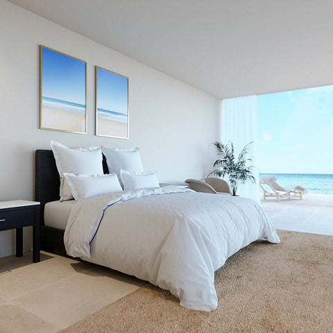 Ein modern hergerichtetes Bett mit einem glatten, weißen Excellence Bettdeckenbezug in einem luxuriösen, hellen Schlafzimmer mit großer Fensterfront und Blick auf den sonnigen Sandstrand.