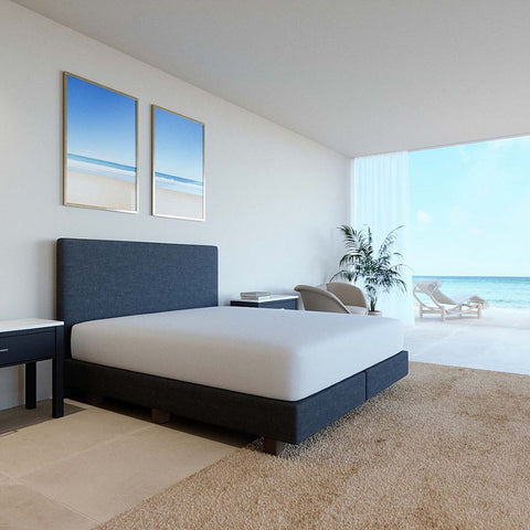 Ein modernes, minimalistisches Boxspringbett mit einem perfekt sitzenden, weißen Premium-Spannbettlaken in einem hellen Schlafzimmer mit großer Fensterfront und Blick auf den Strand.